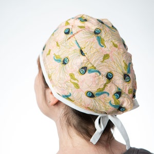 Może przedstawiać: Różowo-biały czepek chirurgiczny z wzorem piór pawia. Czepek ma białe sznurki, które są zawiązane z tyłu głowy.