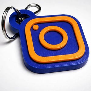 Könnte beinhalten: Ein Schlüsselanhänger mit blauem und orangefarbenem Instagram-Logo. Der Schlüsselanhänger zeigt ein 3D-gedrucktes Design des Instagram-Logos mit blauem quadratischem Hintergrund und orangefarbener Umrandung und Innenkreis. Ein silberner Schlüsselring ist befestigt.