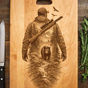 May include: A light-colored wooden cutting board with a handle. The board features an engraved design of a hunter with a rifle, a bear, and a forest scene. Two birds are flying above the hunter.