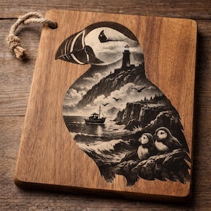 May include: Wooden cutting board with a detailed engraving of a puffin. The puffin's silhouette contains a coastal scene with a lighthouse, boat, and two smaller puffins. The board has a natural wood grain and a rope handle.