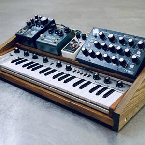 May include: A wooden synthesizer stand with a keyboard and several black and silver effect pedals. The keyboard is labeled "Minimoog Model D (Rack)". The stand is made of light brown wood.