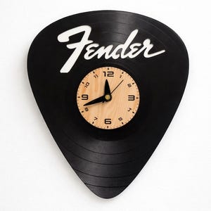 May include: Black wall clock shaped like a guitar pick, with the word "Fender" in white script. The clock face is a light wood color with black hands and numbers.