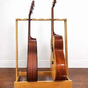May include: A wooden guitar stand holding two acoustic guitars. The stand is made of light-colored wood and has a rectangular base. The guitars are brown and have a glossy finish. The stand is on a wooden floor.