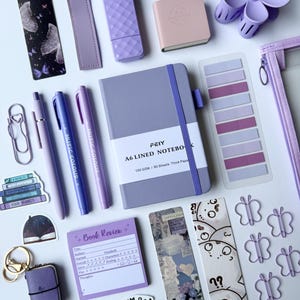 May include: A collection of stationery items in shades of purple and lavender, including an A6 lined notebook, pens, butterfly-shaped paper clips, bookmarks, stickers with book-themed text, a small notepad, and a purple hair claw clip.