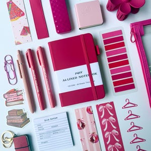 May include: A collection of stationery items in shades of pink and magenta, including an A6 lined notebook, pens, bookmarks with moth and pomegranate patterns, a flower-shaped hair clip, sticky tabs, paper clips, stickers, and a small keychain.