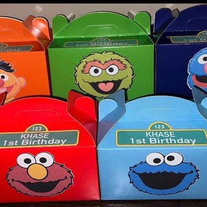 Könnte beinhalten: Eine Sammlung bunter Partygeschenkboxen mit Figuren aus der Sesamstraße. Jede Box hat einen Griff und das Gesicht eines Charakters, darunter Elmo, Krümelmonster und Big Bird. Die Boxen zeigen auch den Text "KHASE 1st Birthday".