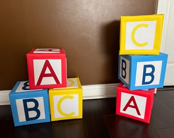 Könnte beinhalten: Bunte Alphabetblöcke in Rot, Gelb und Blau. Jeder Block zeigt einen Buchstaben: A, B oder C. Die Blöcke sind gestapelt und auf einem dunklen Holzboden angeordnet, was eine spielerische und lehrreiche Szene schafft.