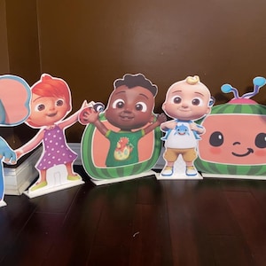 Puede incluir: Una colección de coloridos recortes de cartón independientes con personajes del programa infantil Cocomelon. Los recortes incluyen un elefante, una niña con un vestido morado, un niño en una sandía, un bebé, una sandía y un ratón.