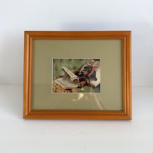 May include: A framed print featuring a dachshund wearing a red polka-dot headscarf and glasses, reclining on a green tufted cushion while reading a newspaper. The artwork is encased in a rectangular wooden frame with a wide, light-colored mat.
