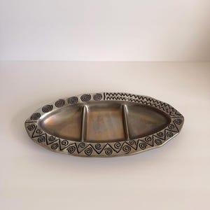 May include: An oval-shaped metal serving tray with three distinct compartments. The rim features a decorative border of etched black spiral and zigzag patterns against a metallic silver-toned surface with a subtle, darkened patina finish.