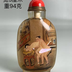 Può includere: Un flacone di vetro trasparente per tabacco da fiuto con tappo rosso e fascia metallica color ottone. La bottiglia presenta una scena dipinta a mano di due figure. Il testo sulla bottiglia recita: 9 cm di altezza, 5 cm di larghezza e pesa 94 grammi.