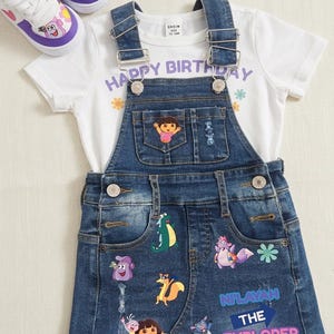 May include: A children's denim overall skirt set with a white t-shirt and matching sneakers. The t-shirt has the words "HAPPY BIRTHDAY" printed on it. The skirt is decorated with cartoon character patches. The sneakers are white and purple.