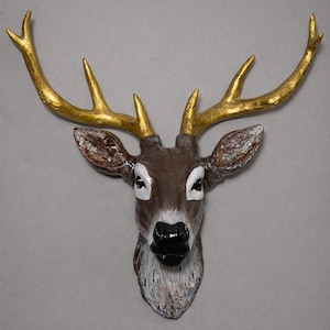 May include: A decorative deer head wall mount with gold-colored antlers. The deer head is painted in shades of brown, white, and black, with a glossy black nose and white accents around the eyes. The antlers are a bright gold color.