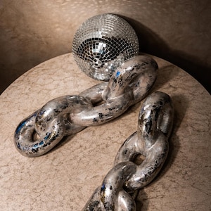 Può includere: Una palla da discoteca argentata e due grandi maglie di catena metalliche poggiano su una superficie rotonda e strutturata. Le maglie della catena hanno un aspetto invecchiato e usurato con sfumature di blu e oro.