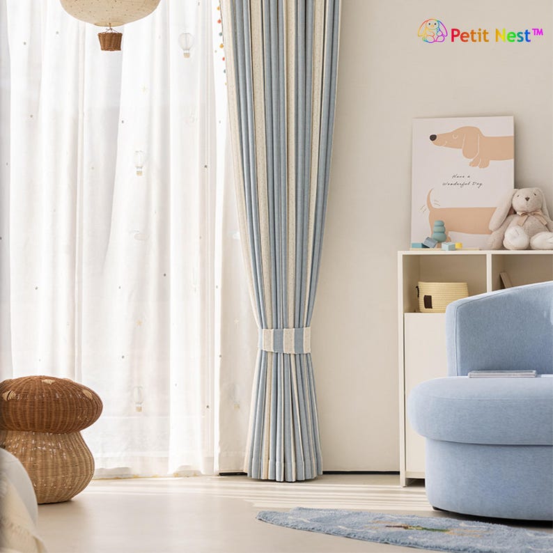 May include: A light blue and white striped curtain is tied back with a matching band. Sheer white curtains with a hot air balloon pattern are visible behind the striped curtain. A light blue armchair and a decorative mushroom-shaped basket are in the foreground.