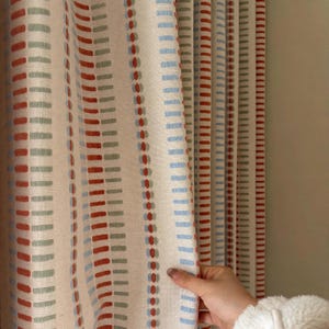 May include: A curtain panel featuring a cream-colored base fabric with vertical stripes composed of repeating dash patterns in shades of burnt orange, sage green, and light blue. The fabric has a visible woven texture and hangs in soft, vertical folds.