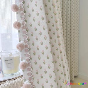 May include: White floral patterned curtain with pink tulip designs and a scalloped edge. The curtain is adorned with pink pom-poms. A candle with the text "AROMATHERAPY NATURAL PLANT" sits on a windowsill.