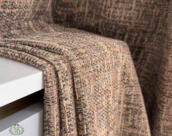 May include: A close-up of a woven fabric in shades of brown, beige, and gray. The fabric is draped over a white surface, showcasing its textured weave. The image highlights the details of the textile, suggesting a home decor item.