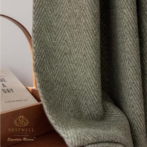 K&ouml;nnte beinhalten: Nahaufnahme eines stoffes in Salbeigr&uuml;n mit Fischgr&auml;tenmuster. Der Stoff ist drapiert und zeigt die Textur und das Gewebe. Das Logo von Nestwell Studio und der Text "Signature Reserve" sind im unteren Teil des Bildes sichtbar. Ein Buch ist teilweise im Hintergrund zu sehen.