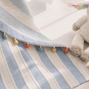 May include: A blue and white striped textile with a decorative edge featuring small, colorful pom-poms. The fabric has a textured appearance and a subtle herringbone weave. The overall design is simple and elegant, suitable for home decor.