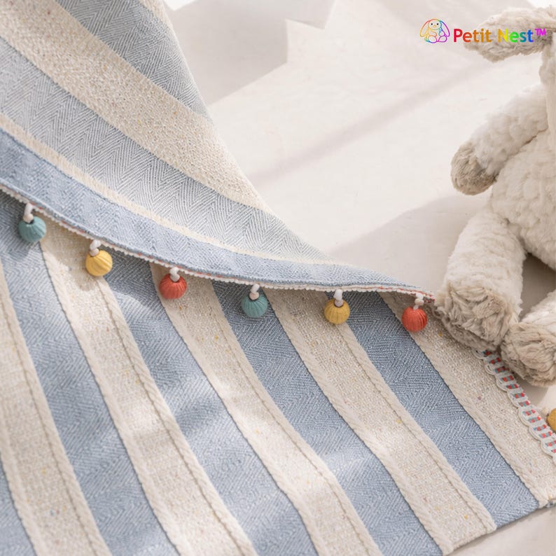 May include: A blue and white striped textile with a decorative edge featuring small, colorful pom-poms. The fabric has a textured appearance and a subtle herringbone weave. The overall design is simple and elegant, suitable for home decor.