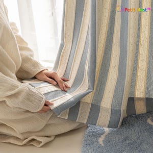 May include: Blue and cream striped curtain with a textured weave. The curtain is held by a person wearing a cream-colored sweater. The curtain is hanging in front of a window, with a blue rug visible in the foreground.