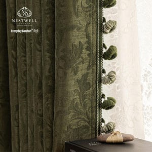 May include: Olive green patterned curtain with a floral design. The curtain has a decorative trim with green and white tassels. The text "NESTWELL STUDIO" and "Everyday Comfort" is visible on the curtain.