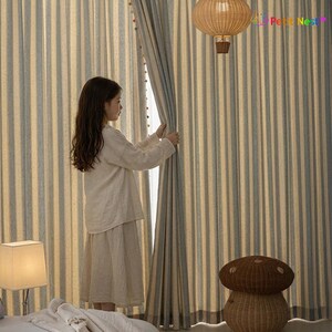 May include: A young person stands near striped curtains, opening them to reveal a bright space. The curtains have vertical stripes in light blue and beige. A woven mushroom-shaped basket and a rattan pendant light are also visible.