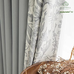May include: Gray textured curtains next to floral print curtains. The floral curtains have a white background with gray botanical illustrations. The text "NESTWELL STUDIO Everyday Comfort" is visible on the floral curtain.