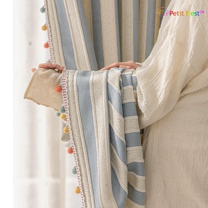 May include: Striped curtain panels in light blue and cream with a textured weave. The edges are trimmed with a delicate lace and colorful pom-poms. The curtains are hanging in front of a window.