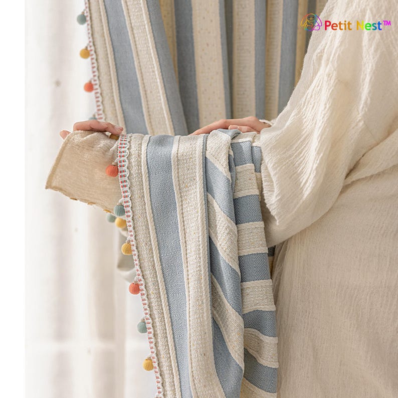 May include: Striped curtain panels in light blue and cream with a textured weave. The edges are trimmed with a delicate lace and colorful pom-poms. The curtains are hanging in front of a window.