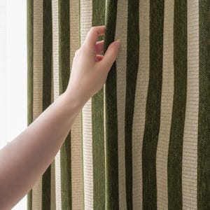 May include: A curtain featuring a vertical striped pattern in alternating shades of dark forest green and light beige. The fabric has a visible woven texture, and the stripes run parallel from top to bottom, creating a consistent linear design.