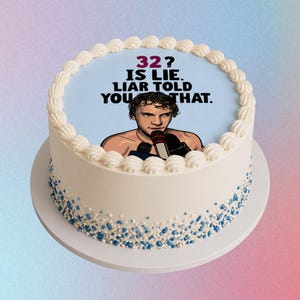 May include: A white frosted cake with blue and white sprinkles around the base. The top of the cake has a light blue background with the text "32? IS LIE. LIAR TOLD YOU THAT." and a cartoon image of a person.