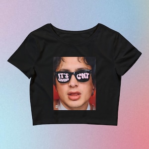 Puede incluir: Top corto negro con un gráfico de una persona con gafas de sol. Las gafas de sol tienen el texto "IT'S GIVING CNT" en rosa. La imagen es un primer plano de la cara de la persona. El top es de estilo casual.