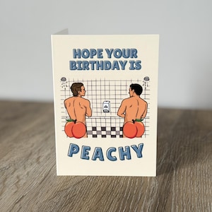 May include: Greeting card with the text "HOPE YOUR BIRTHDAY IS PEACHY". The card features a cartoon illustration of two men in a shower with peach-shaped bottoms. The background is a grid pattern.