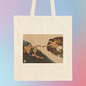 May include: A cream-coloured canvas tote bag featuring a printed graphic of two figures reaching towards each other. The figure on the left wears blue shorts, while the figure on the right wears white shorts, both set against a tan background.