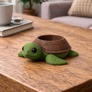 May include: A turtle-shaped planter with a textured green head and flippers, featuring a brown, segmented shell that forms a circular opening. The object has large, smooth black eyes and a detailed, ridged surface texture throughout its body.