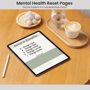 May include: A digital tablet with "Reset & Breathe" prompts, a white stylus, a glass of water, a coffee cup, and a plate of white treats. The text "Mental Health Reset Pages" is at the top.