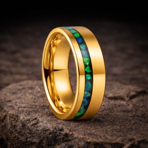 May include: A gold-colored ring with a central inlay of green and blue opal-like material. The ring is set against a dark, textured background, highlighting its polished surface and gemstone detail.