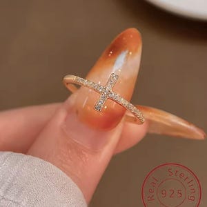 Peut inclure: Une bague dorée avec un motif de croix. La croix et le bandeau sont incrustés de petits pierres précieuses transparentes. La bague est tenue par le doigt d'une personne. La bague est estampillée "Real Sterling 925 Silver".