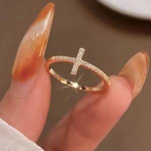 May include: A gold ring with a cross design. The ring's band is set with small, sparkling stones, and the cross is also decorated with similar stones. The ring is held up by a person's fingers.