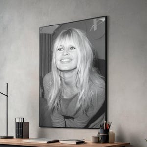 May include: A black and white framed wall art print featuring a portrait of a person with long blonde hair, bangs, and a smiling expression. The subject is wearing a long-sleeved top and is captured in a medium shot, displayed in a thin black frame.