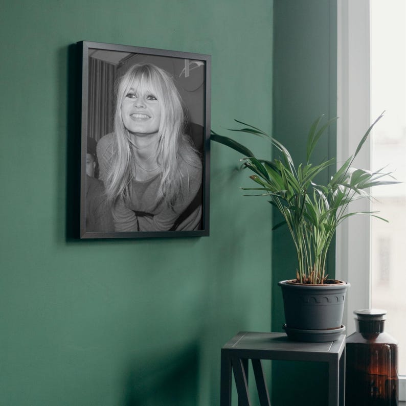 May include: A black and white framed photograph of a person with long blonde hair and bangs, smiling. The portrait is displayed in a thin black frame hanging on a solid green wall, positioned above a dark gray side table with a green potted plant.