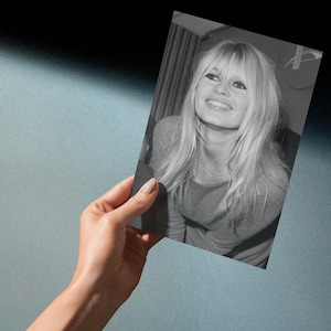 May include: A black and white rectangular photograph featuring a portrait of a smiling person with long blonde hair, bangs, and dark eyeliner. The person is wearing a long-sleeved top and a thin necklace, captured in a medium shot with a soft expression.