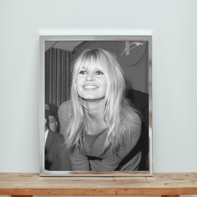 May include: A black and white framed photograph featuring a portrait of a person with long blonde hair, bangs, and a smiling expression. The subject is wearing a long-sleeved top and is positioned in front of a partially visible person in a suit.