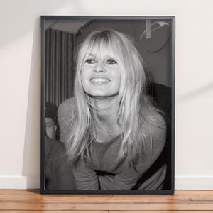 May include: A black and white framed photograph featuring a portrait of a smiling individual with long, layered blonde hair and bangs. The subject is wearing a long-sleeved top and a thin necklace, captured in a candid, upward-looking expression.