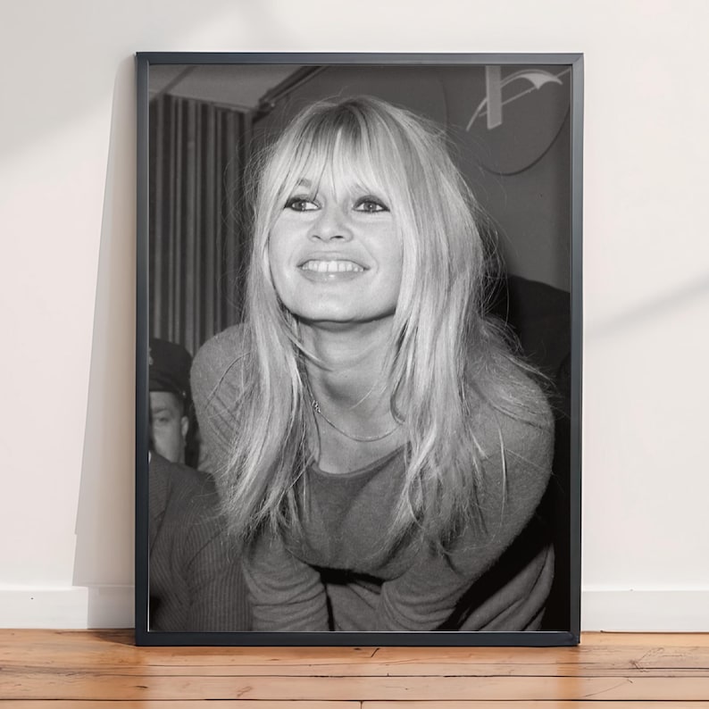 May include: A black and white framed photograph featuring a portrait of a smiling individual with long, layered blonde hair and bangs. The subject is wearing a long-sleeved top and a thin necklace, captured in a candid, upward-looking expression.