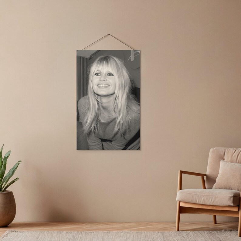 May include: A black and white rectangular wall hanging featuring a portrait of a smiling individual with long, light-colored hair and bangs. The portrait is suspended by a thin string attached to the top, displaying a clear, high-contrast image.