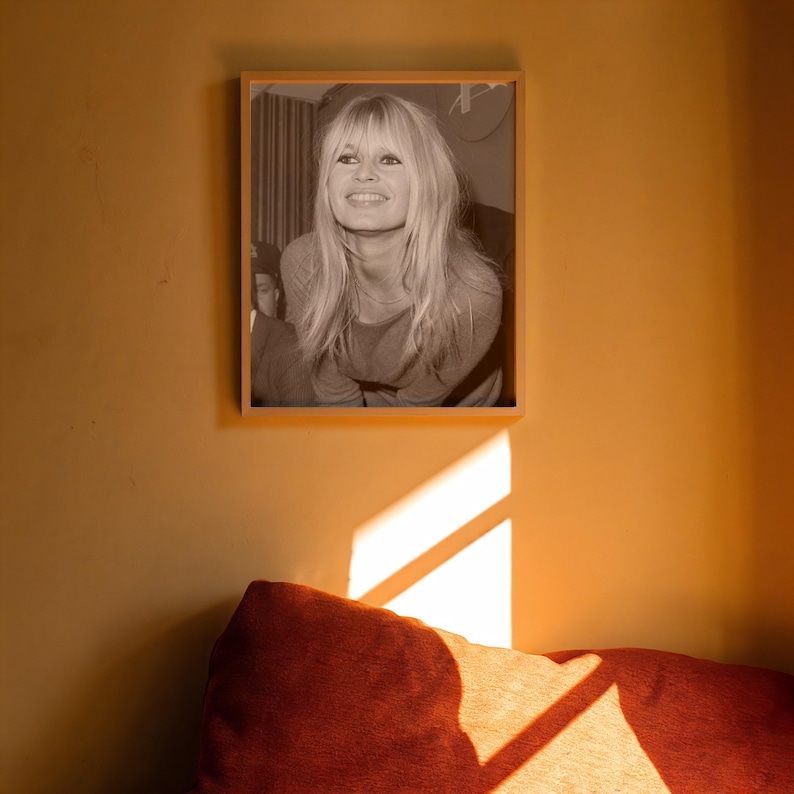 May include: A rectangular framed black and white photograph featuring a portrait of a smiling person with long blonde hair. The print is mounted in a light-colored wooden frame and is displayed on a solid, warm-toned mustard yellow wall.