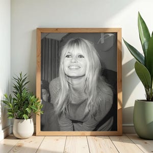 May include: A black and white framed photograph of a person with long blonde hair and bangs, smiling while looking upward. The person wears a long-sleeved top and a thin necklace. The portrait is enclosed in a light-colored wooden frame.